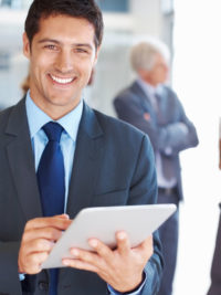 Portrait of smiling young business man working on digital tablet with executives in background